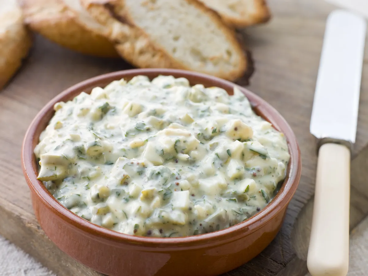 Un bol de sauce gribiche garnie d'œufs durs hachés, câpres et herbes fraîches posé sur une planche en bois