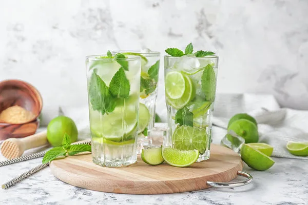 Verre transparent de mojito sans alcool garni de feuilles de menthe, quartiers de citron vert et glace pilée posé sur une table en bois clair
