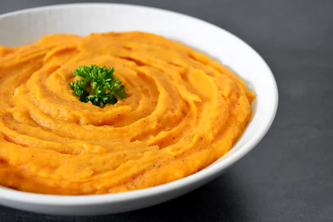 Bol en céramique rempli d'une purée orange crémeuse, garni de feuilles de coriandre et de graines, posé sur une table en bois clair