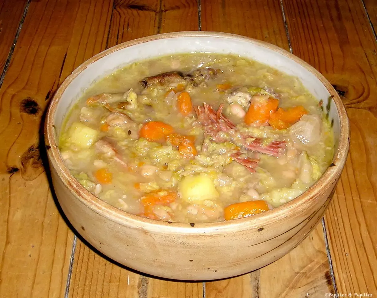 Bol fumant de garbure posé sur une table en bois, morceaux de chou, carottes, haricots blancs et une cuillère en bois