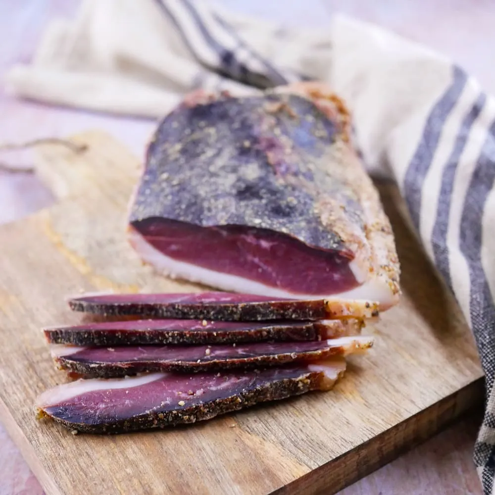 Deux tranches fines de magret séché posées sur une planche en bois, autour quelques grains de poivre et brins de thym, lumière douce du matin