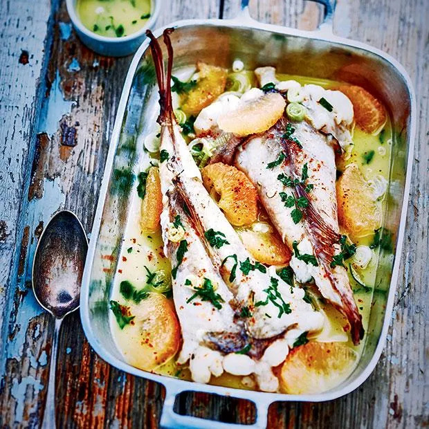 Queue de lotte rôtie dans une cocotte en fonte, entourée de tomates cerises et d'herbes fraîches, sur une table en bois brut éclairée par la lumière naturelle