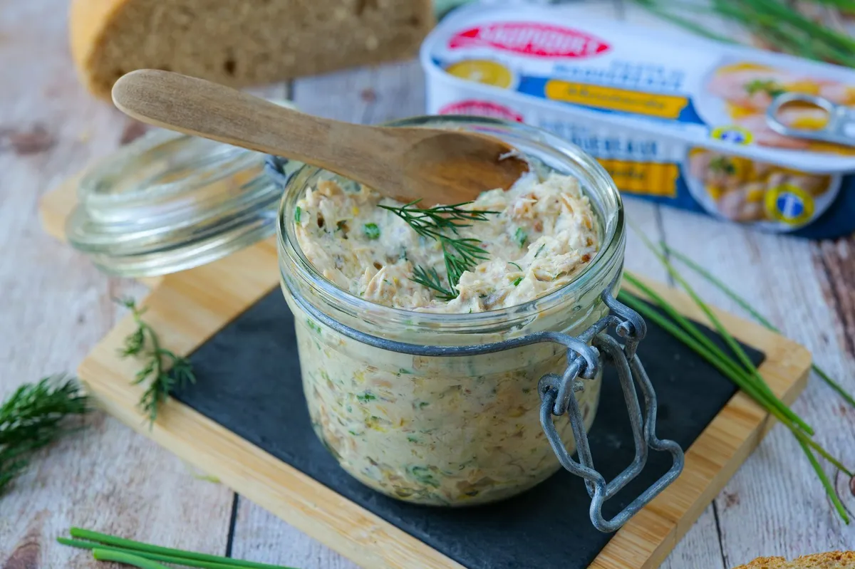 Un pot en verre ouvert contenant des rillettes beige clair, morceaux de maquereau visibles, herbes fraîches, quartiers de citron et pain grillé sur une table en bois.
