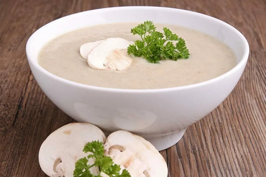 Un bol rustique rempli d'un velouté beige crémeux, garni de persil ciselé, posé sur une table en bois avec une tranche de pain grillé à côté